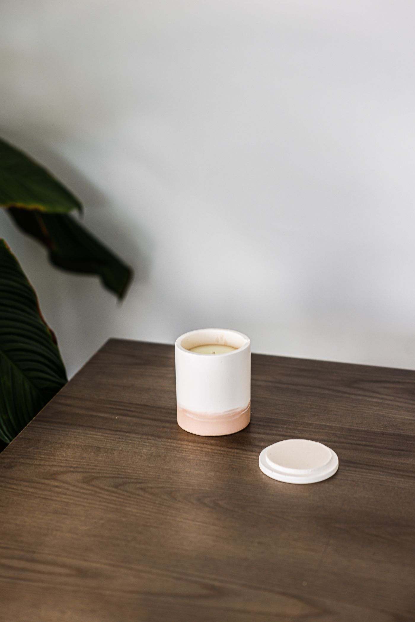 A cylindrical ceramic candle jar, white above and blush pink on about the bottom third, open, with pale wax inside. The separate lid is pale blush with a white rim. It sits on a dark brown wooden table, with green leaves entering from the left. Photograph by Cup of Couple. Not client work.