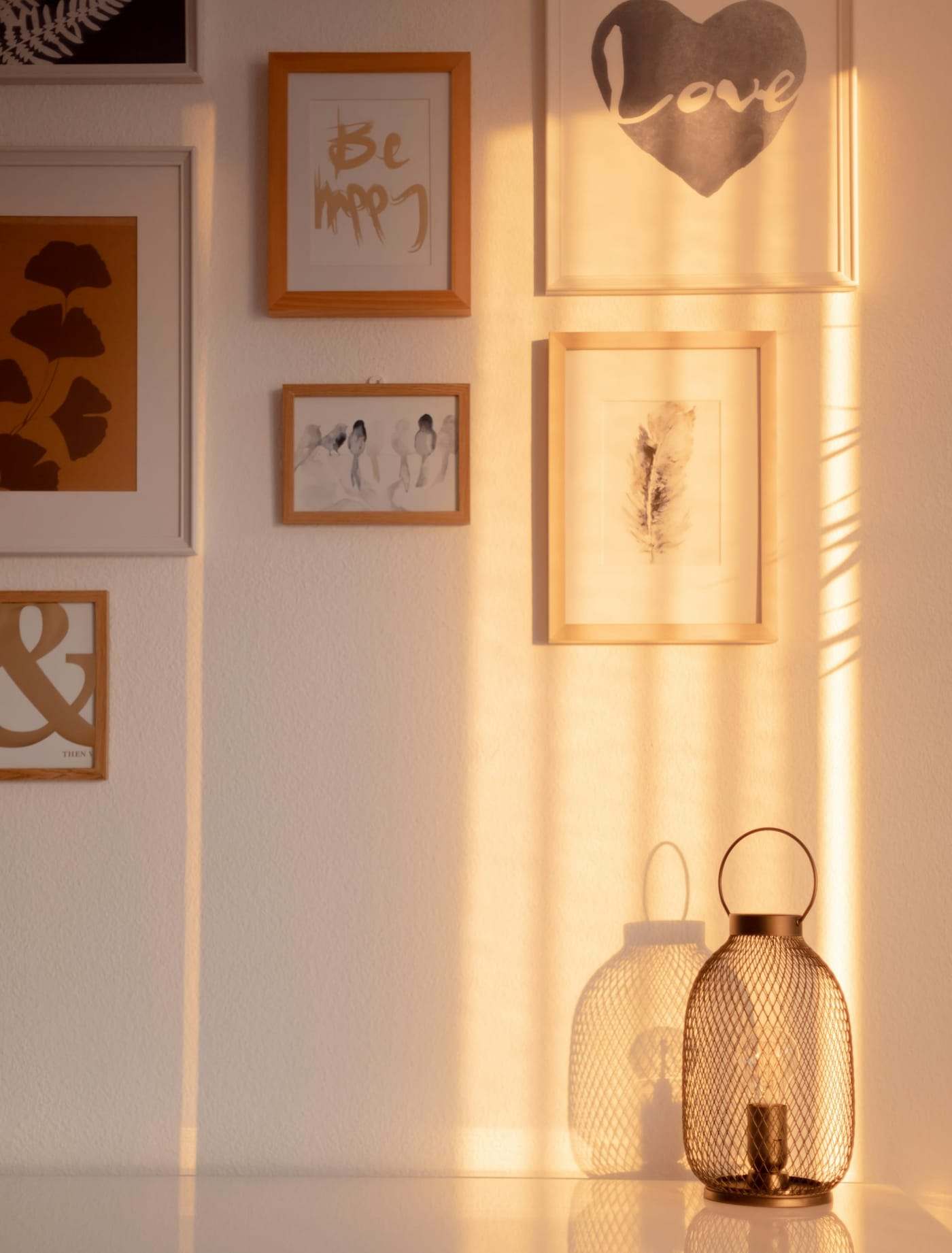 A bronze diamond-mesh lantern with a ring handle and a clear bulb, sitting on a glossy white surface against a cream wall hung with framed prints, in warm bands of sunlight. Photograph by Ryan Klaus. Not client work.