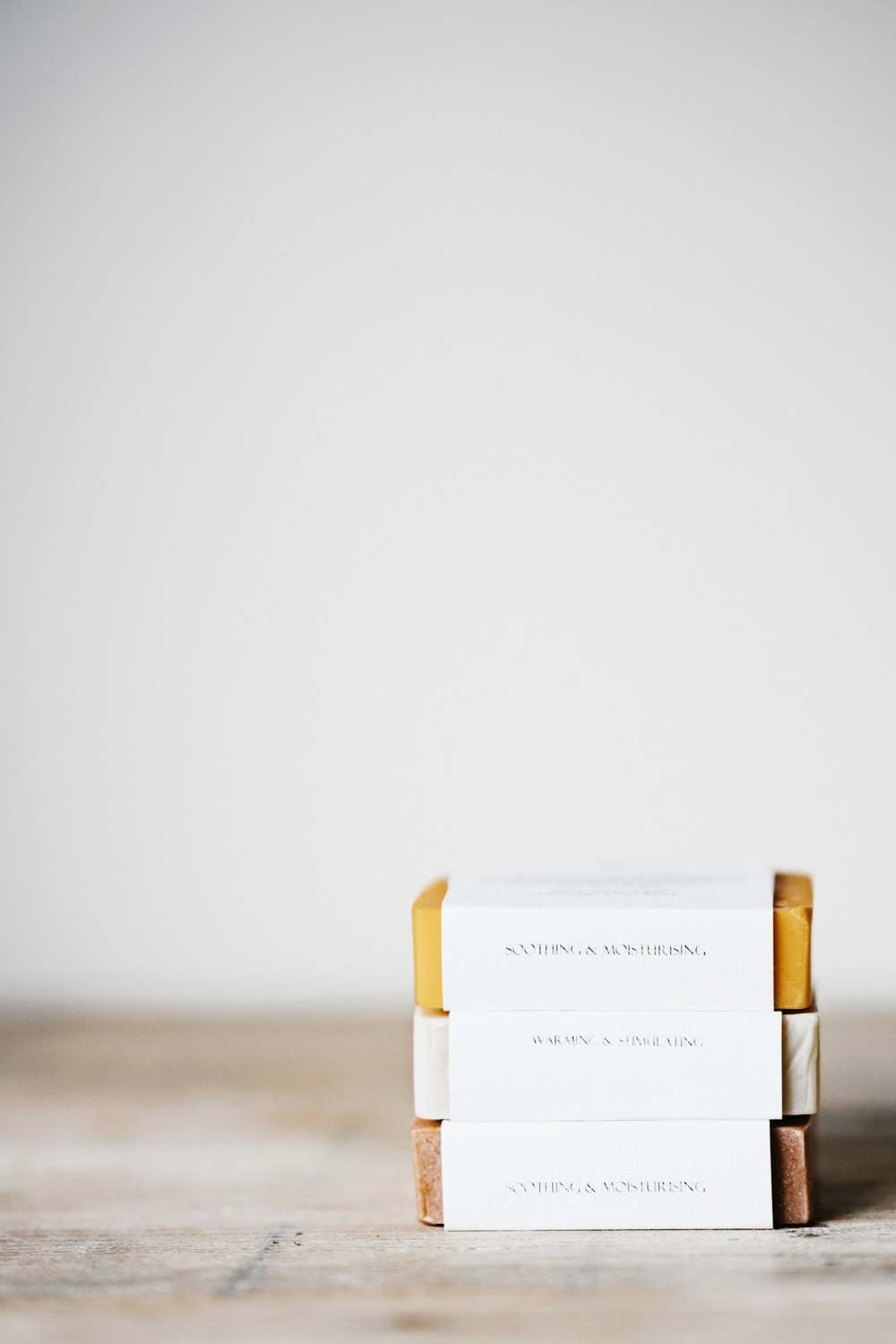Three stacked soap bars on a pale wooden surface, against a near-white wall. The top bar is mustard yellow, the middle bar is cream, and the bottom bar is speckled brown. Each has a white paper band. Photograph by Annie Spratt. Not client work.